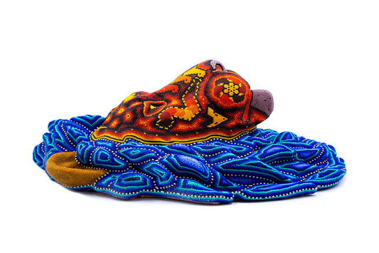Pieza artesanal representandola cabeza de un leon, con una compleja y colorida decoración de cuentas que sigue el estilo Huichol. La figura se muestra en tonos cálidos como el naranja, rojo y amarillo, con toques de azul, reposando sobre una base ondulada de motivos azules y geométricos