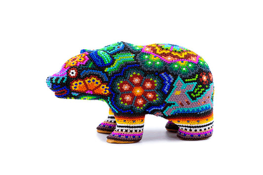 Vista lateral de figura de oso con decoración Huichol en cuentas, con colores brillantes y variados que forman patrones tradicionales, destacando sobre un fondo neutro