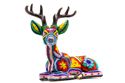 Vista frontal de una escultura de venado acostado, cubierta con chaquiras en un mosaico de colores brillantes que forman diseños tradicionales huicholes. El venado presenta una expresión serena y cuernos detallados, reflejando la artesanía exquisita