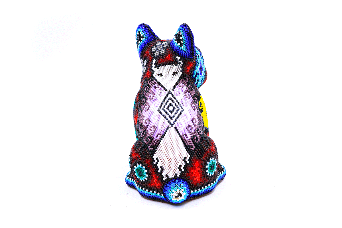 Artesanía Huichol representando un perro bulldog sentado, meticulosamente adornado con cuentas coloridas en patrones Wixárika, destacando en un fondo blanco.