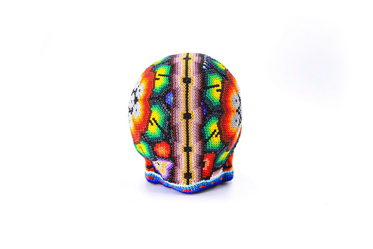 Artesanía huichol en forma de cráneo vibrante, minuciosamente decorado con un mosaico de cuentas de colores que forman patrones tradicionales wixárika, resaltando tonos predominantes en naranja, verde y amarillo sobre fondo blanco