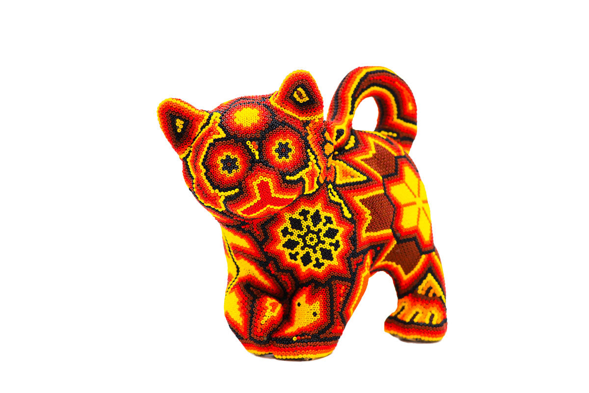 Artesanía huichol en forma de gato de frente, decorada con un patrón detallado de chaquiras en tonos rojos, amarillos y naranjas, destacando sobre un fondo blanco. El diseño incluye flores y símbolos geométricos que reflejan la rica tradición y simbolismo de la cultura wixárika.