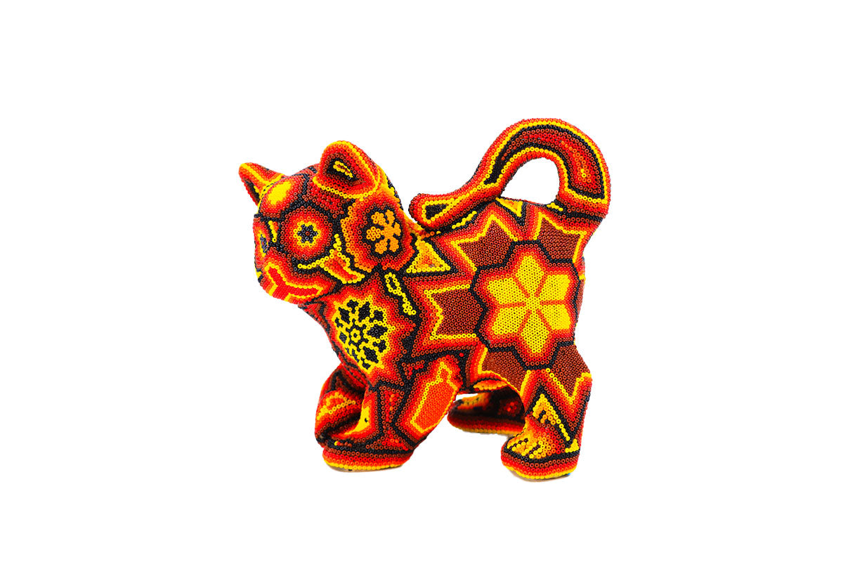 Artesanía huichol en forma de gato de perfil, decorada con un patrón detallado de chaquiras en tonos rojos, amarillos y naranjas, destacando sobre un fondo blanco. El diseño incluye flores y símbolos geométricos que reflejan la rica tradición y simbolismo de la cultura wixárika.