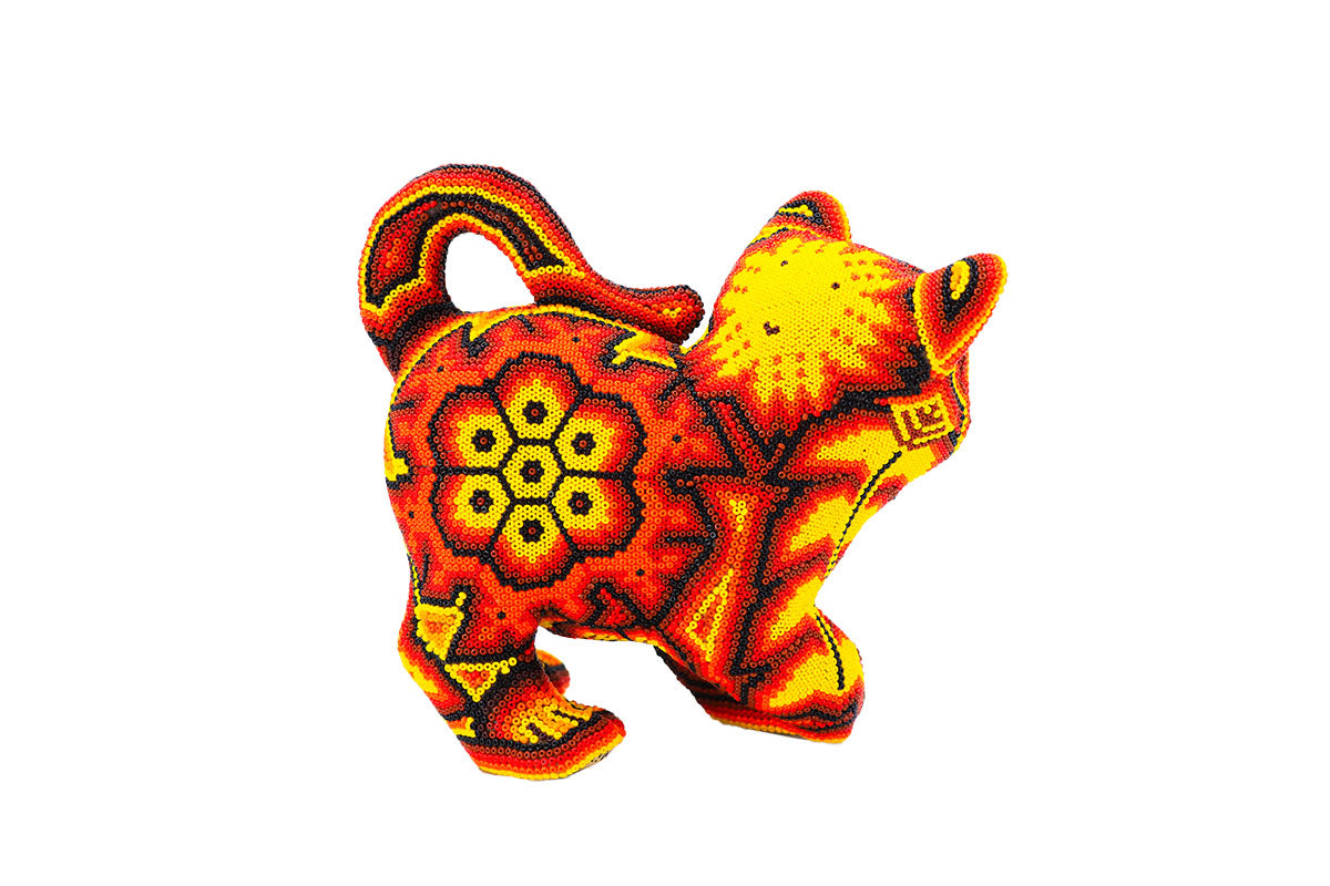 Artesanía huichol en forma de gato de espalda, decorada con un patrón detallado de chaquiras en tonos rojos, amarillos y naranjas, destacando sobre un fondo blanco. El diseño incluye flores y símbolos geométricos que reflejan la rica tradición y simbolismo de la cultura wixárika.