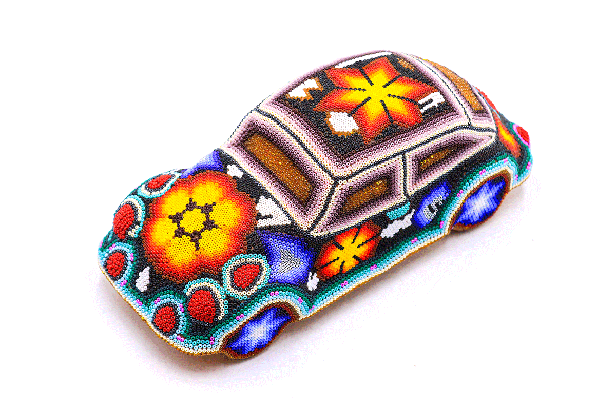 Vista superior de una artesanía de un coche huichol, con un diseño de chaquiras multicolores creando flores y formas simbólicas, sobre un fondo que combina colores vivos como el azul, rojo y amarillo