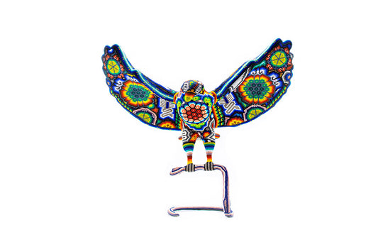 Figura artesanal de águila con alas extendidas, elaborada con cuentas de colores vivos siguiendo patrones tradicionales Huichol, mostrando una serpiente en su pico, sobre un fondo blanco