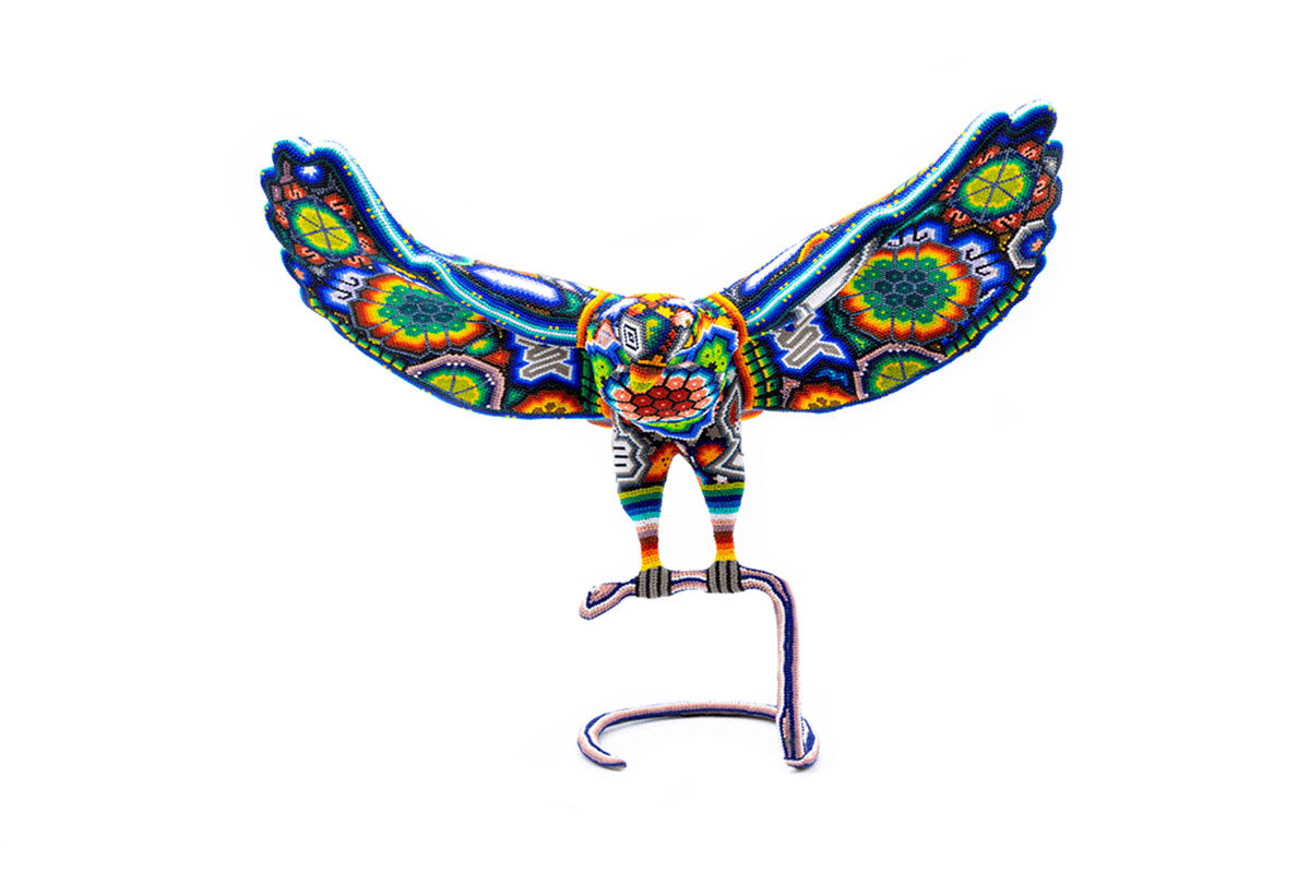 Artesanía de un águila en posición de ataque con serpiente en boca, con detalle de cuentas en colores primarios y patrones Huichol, posicionada sobre una base de metal, contra un fondo blanco