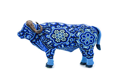 Artesanía en forma de búfalo adornada con intrincados patrones azules y blancos en estilo Huichol, utilizando cuentas pequeñas para crear un mosaico detallado. El animal está en posición de pie con cuernos curvos y realistas que contrastan con el vibrante trabajo de chaquira