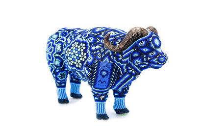 Escultura de búfalo decorada con chaquiras en tonos de azul y blanco, mostrando patrones geométricos y florales característicos del arte Huichol. La pieza de artesanía resalta por su precisión en los detalles y la armonía de sus colores, con cuernos marrones texturizados que complementan su diseño estético