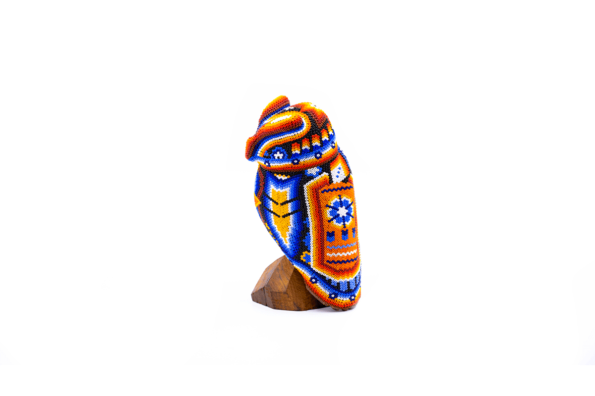 Escultura de búho huichol de tamaño pequeño sobre base de madera, con intrincado patrón de cuentas multicolores. Destacan los tonos vibrantes de azul, naranja y amarillo, con detalles como estrellas y formas geométricas típicas del arte huichol. La pieza refleja la rica tradición artesanal y espiritual de los huicholes de México
