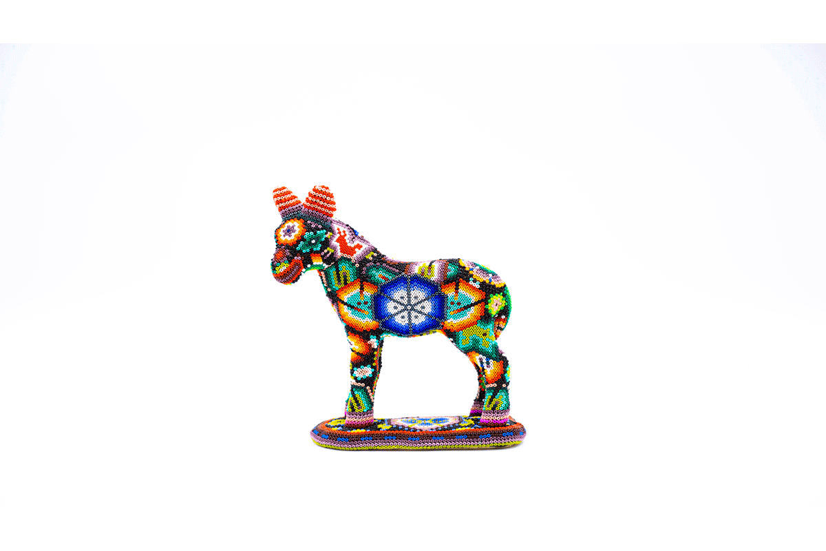 Figura artesanal de un burro en posición de pie, ricamente decorada con chaquiras multicolores en patrones complejos y simétricos de arte huichol, destacando figuras geométricas y estrellas sobre un fondo blanco