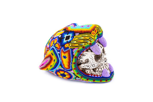 Artesanía de una cabeza de jaguar hecha con chaquira, cubierta con un patrón de arte Huichol vibrante que mezcla púrpuras, amarillos y azules, con la boca abierta mostrando dientes tallados en detalle en su interio