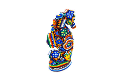igura artesanal en forma de caballito de mar de perfil, decorada con intrincados patrones de chaquira en colores vibrantes como azul, naranja, verde y amarillo, sobre fondo blanco