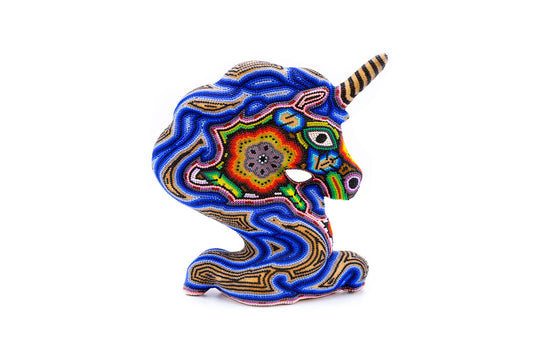 Escultura artesanal en forma de cabeza de unicornio, perfilada con cuentas de chaquira. Destacan los patrones ondulados y geométricos en colores azul, rojo y verde sobre un fondo blanco