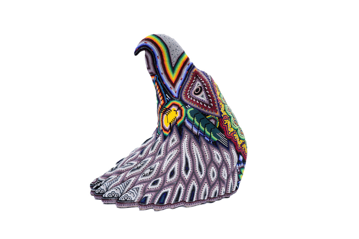 Escultura de cabeza de águila huichol meticulosamente embellecida con un patrón de chaquiras de colores vibrantes, destacando sobre un fondo blanco. El diseño incluye elementos simbólicos tradicionales wixárikas, como flores y estrellas, en una gama de colores que incluye morados, grises, amarillos y azules