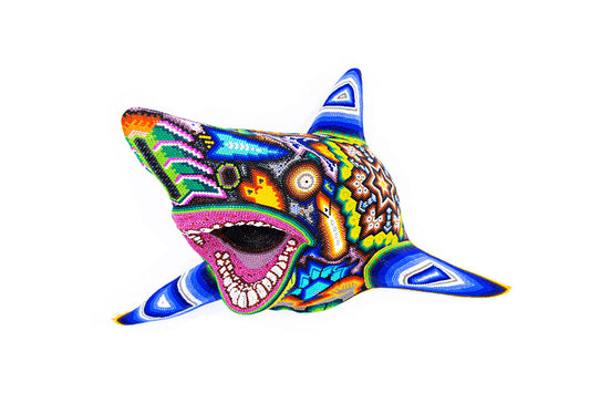 Figura decorativa de una cabeza de tiburón vista de frente, caracterizada por su mosaico de chaquiras que crean una expresión vibrante, con un predominio de azules, amarillos y rojos, sobre un fondo blanco puro