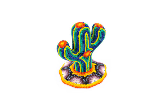 Escultura artesanal de un cactus pequeño con tres brazos, cubierta de chaquiras en colores brillantes como azul, naranja y verde, destacando sobre un suelo simulado con cuentas en tonos marrones y amarillos, sobre un fondo blanco