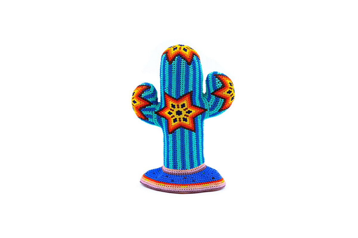 Escultura artesanal de un cactus alto con dos brazos, cubierta por chaquiras que forman diseños geométricos y patrones tribales en tonos de azul, naranja y amarillo, con detalles en rosa y verde, sobre un fondo blanco