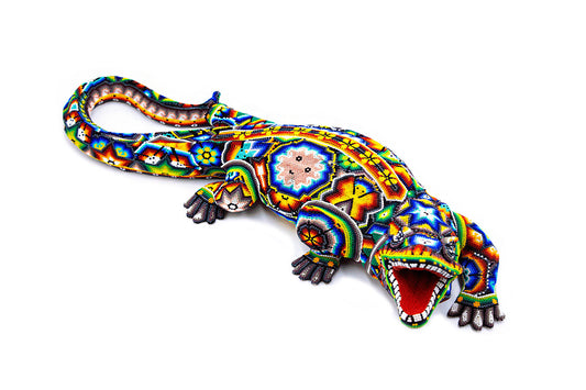 Escultura artesanal de un cocodrilo elaborada con la técnica de chaquira, presentando un diseño vibrante con patrones tradicionales Huichol en una gama de colores brillantes como azul, amarillo, rojo y verde, con la boca abierta mostrando una lengua roja y dientes blancos