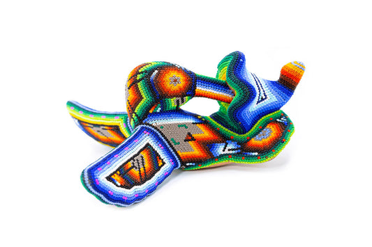 Figura artesanal de un colibrí en posición de vuelo, adornado con un patrón de chaquiras multicolor. Se observan combinaciones de naranja, azul, y verde, con detalles en blanco sobre fondo neutro
