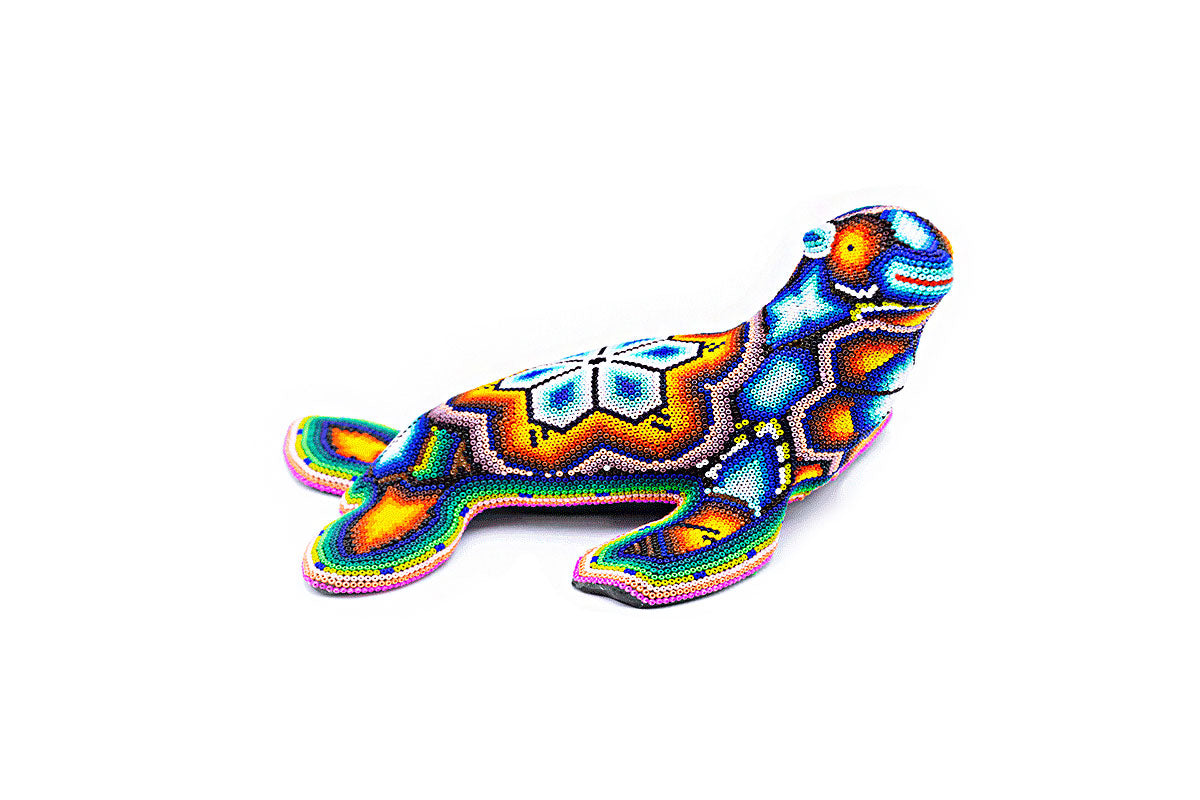 Figura artesanal de una foca en posición recostada, decorada con minucioso trabajo de chaquira en colores brillantes y patrones Huichol, destacando sobre un fondo blanco