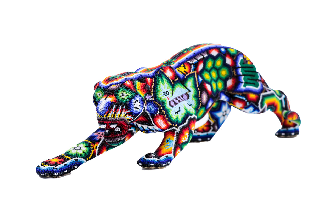 Escultura de jaguar huichol en posición de acecho, ricamente decorada con patrones de chaquiras que representan la iconografía wixárika, sobre un fondo blanco. La paleta de colores incluye tonos vibrantes de azul, verde, rojo, enfatizando la destreza artesanal de la cultura huichol