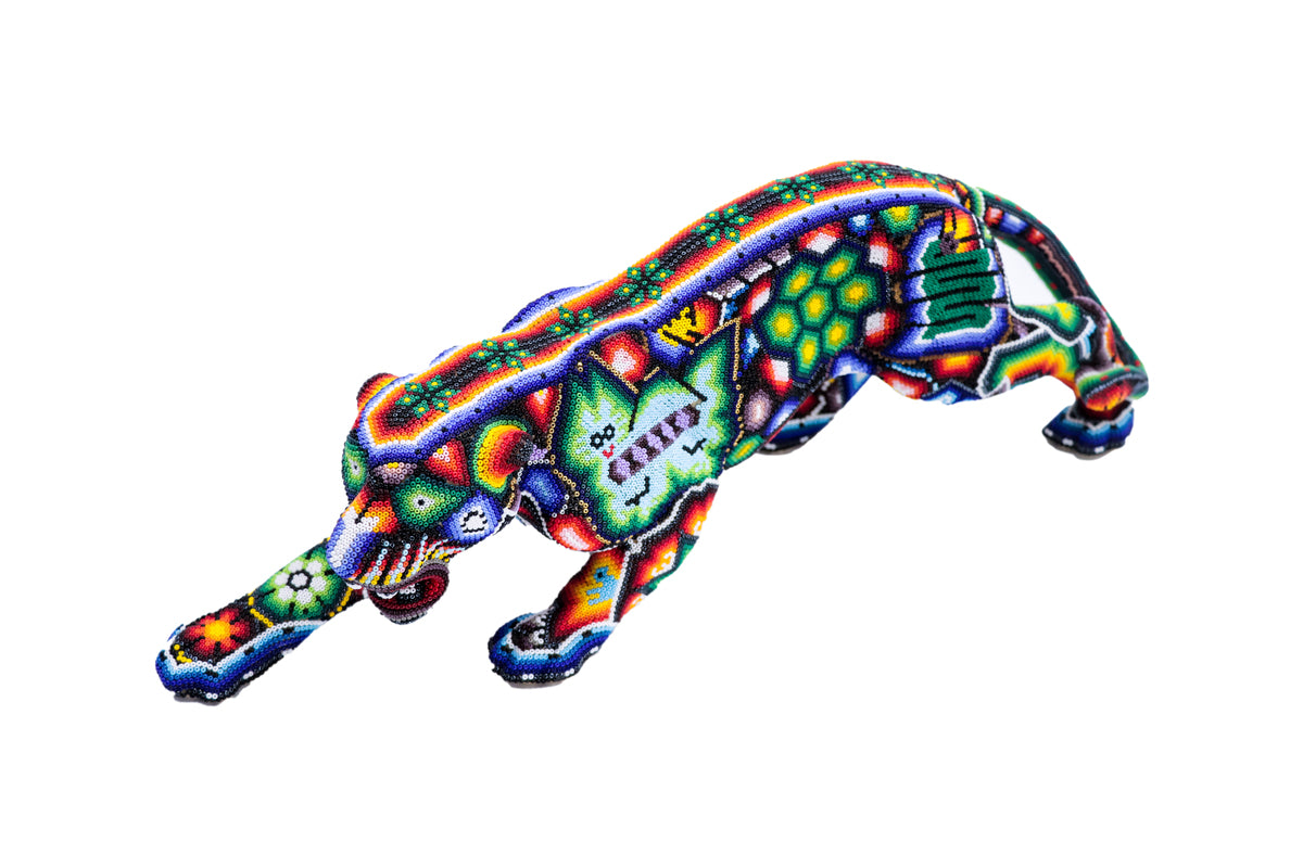 Escultura de jaguar huichol en posición de acecho cenital, ricamente decorada con patrones de chaquiras que representan la iconografía wixárika, sobre un fondo blanco. La paleta de colores incluye tonos vibrantes de azul, verde, rojo, enfatizando la destreza artesanal de la cultura huichol