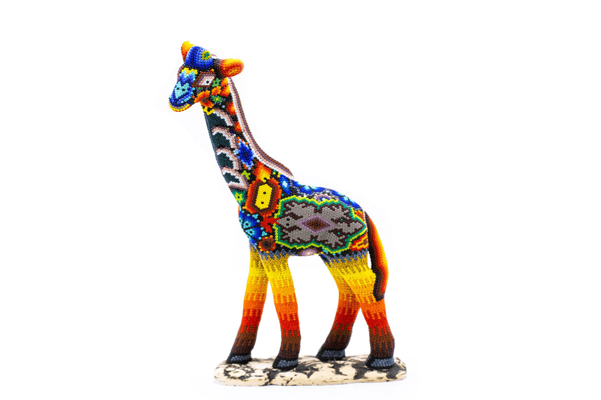 Escultura artesanal en forma de jirafa, cubierta con un mosaico de cuentas brillantes y coloridas. Presenta un patrón vibrante con predominio de tonos azules en el cuello y amarillos, naranjas y rojos en las patas. La base imita las manchas de la jirafa con un diseño de mosaico
