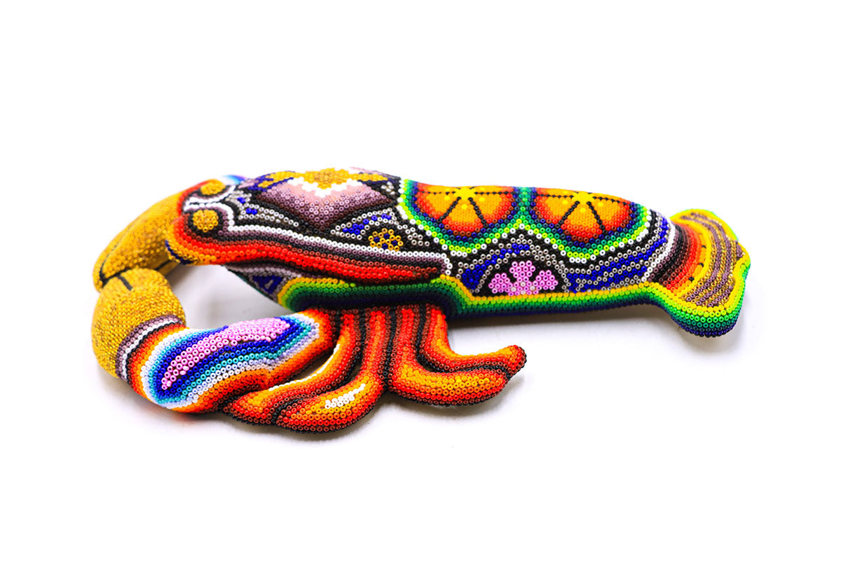 Escultura artesanal de una langosta en estilo huichol, cubierta con un mosaico de chaquiras de colores brillantes. El diseño cuenta con patrones geométricos y símbolos tradicionales huicholes en una gama vibrante de colores como amarillo, naranja, verde y azul, con detalles en blanco y negro