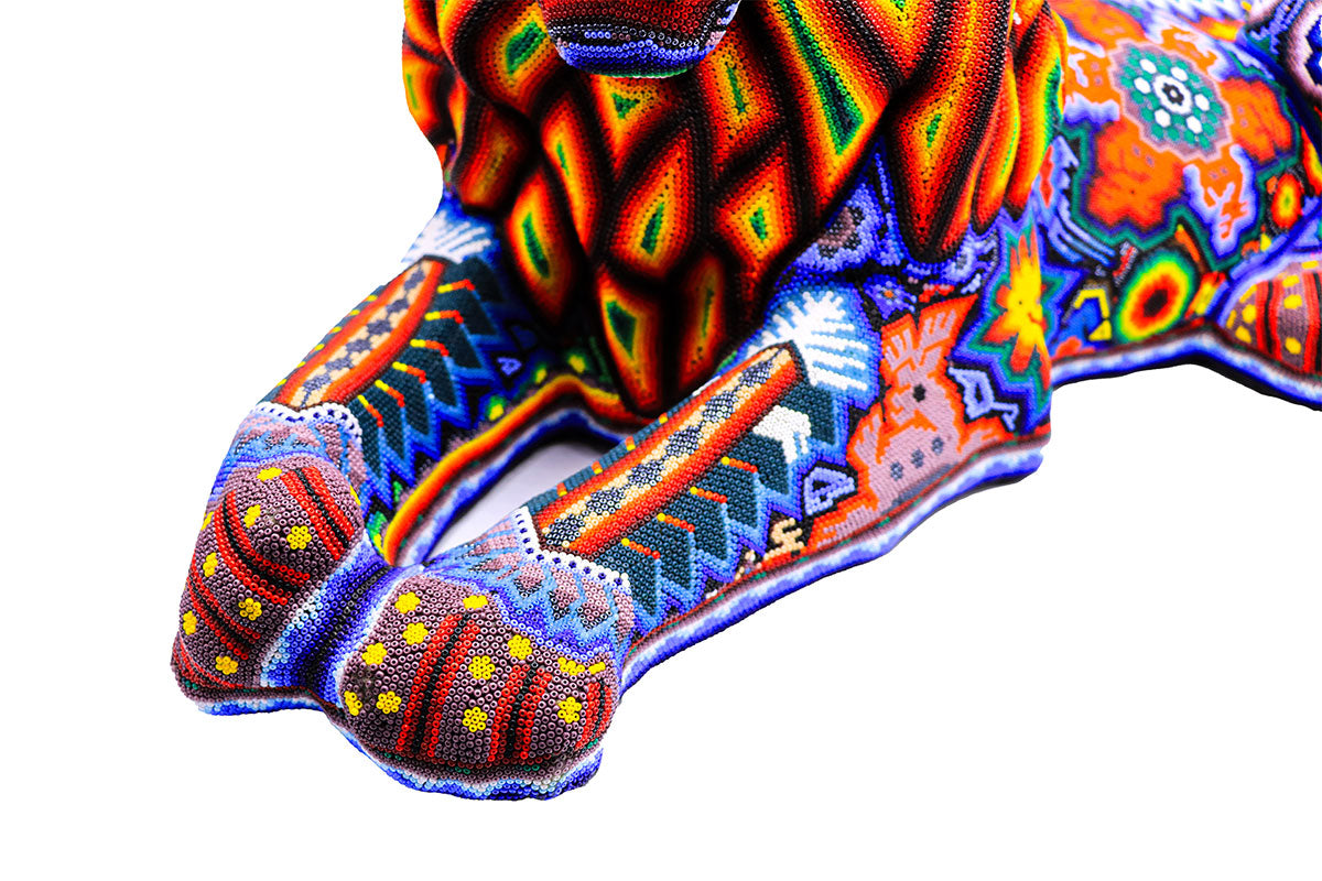 Obra de arte Huichol en forma de león acostado, decorada con cuentas brillantes y diseño psicodélico, expresando la cosmovisión y el arte tradicional del pueblo Wixárika