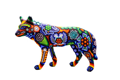 Artesanía huichol en forma de un lobo, cubierto completamente con un mosaico de chaquiras que forman diseños tradicionales Wixarika, tales como mandalas, estrellas y flores de peyote, en una fusión de colores brillantes como el azul, rojo, naranja y verde, contra un fondo blanco que resalta la riqueza visual de la pieza
