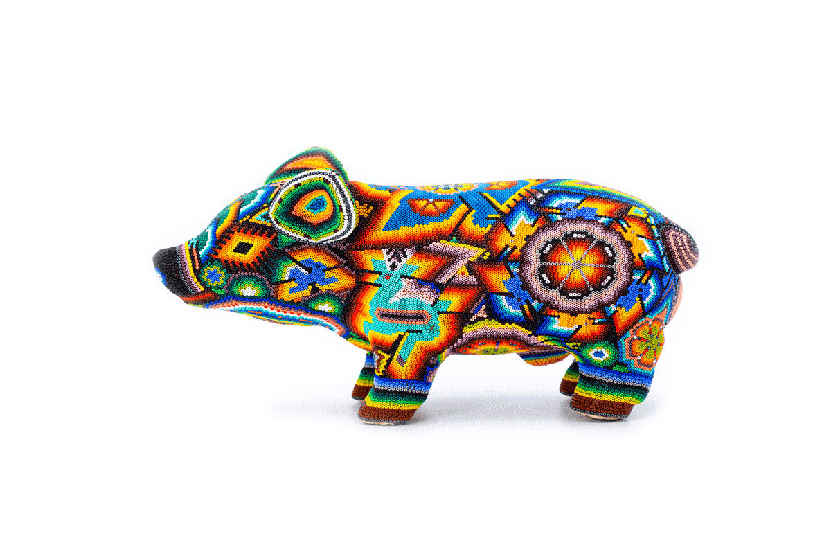 Figura artesanal de un cerdo en vista lateral, cubierto por un mosaico de cuentas coloridas en un estilo Huichol distintivo, que presenta patrones geométricos y simbólicos en tonos brillantes de rojo, azul, verde y amarillo, con una flor prominente en su lomo.