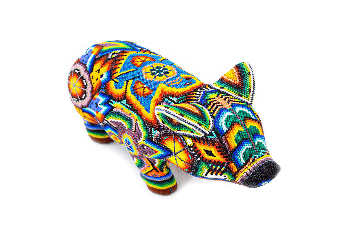 Escultura de un cerdo Huichol vista desde arriba, exhibiendo un tapiz de intrincados patrones de chaquira con una gama de colores vivos como azul, naranja, y verde, con un diseño complejo que incluye figuras geométricas y elementos de la naturaleza.