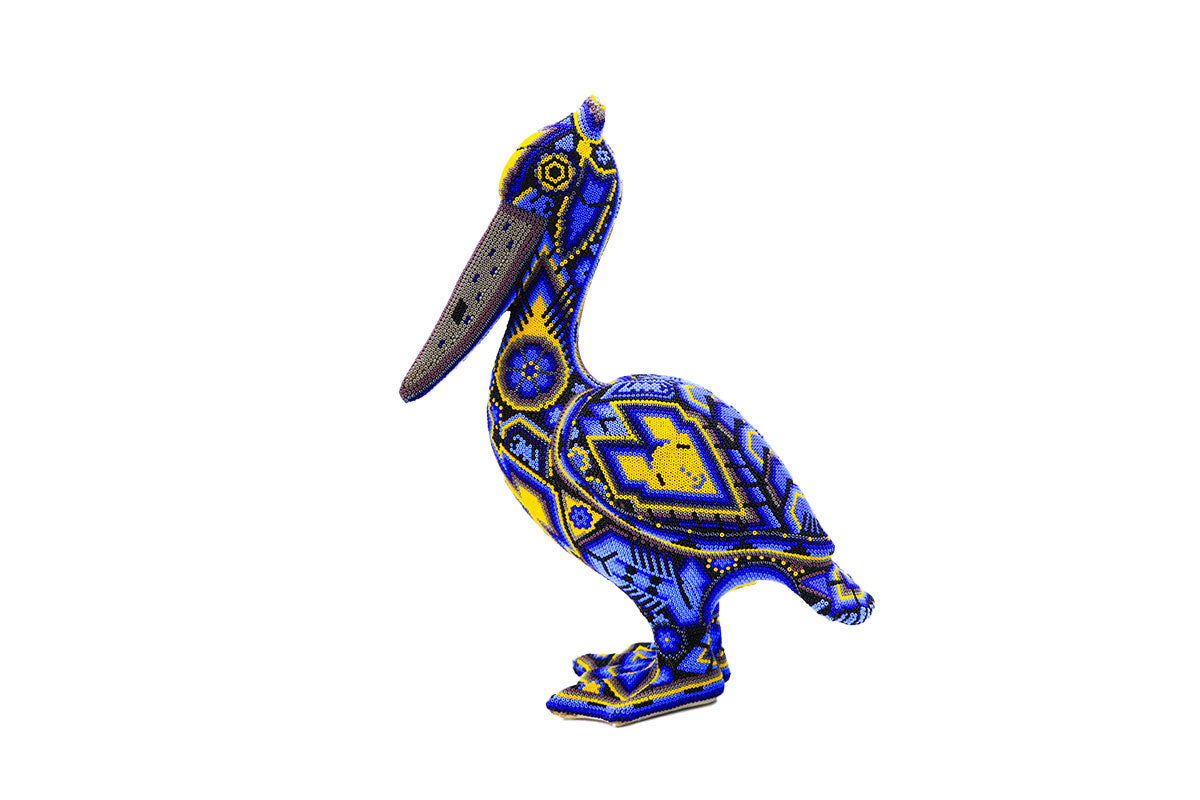 Escultura de pelícano huichol de pie y perfil, cubierta completamente con chaquiras en un vibrante patrón de azules y amarillos que forman diseños geométricos y simbólicos tradicionales wixárikas. La artesanía detallada se destaca poderosamente contra un fondo blanco.
