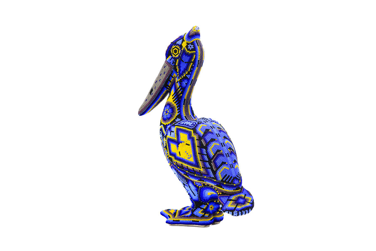 Escultura de pelícano huichol de pie, cubierta completamente con chaquiras en un vibrante patrón de azules y amarillos que forman diseños geométricos y simbólicos tradicionales wixárikas. La artesanía detallada se destaca poderosamente contra un fondo blanco