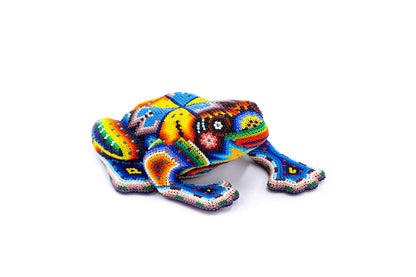 Artesanía de una rana en posición reclinada, adornada con chaquiras de colores vivos que forman patrones abstractos y florales. Resaltan los tonos azules, amarillos y rojos sobre un fondo blanco