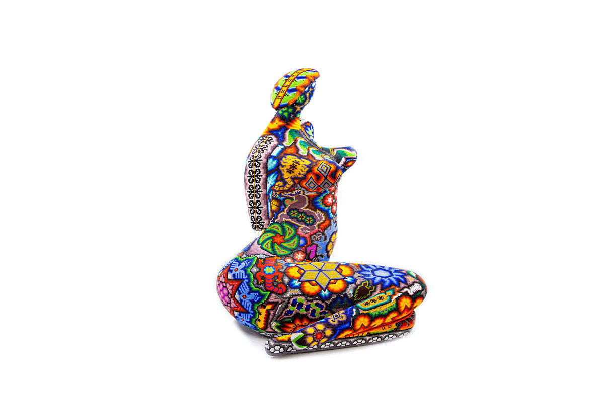 Escultura artesanal de un torso de mujer, decorado con patrones vibrantes y coloridos en estilo Huichol. La figura muestra una mezcla de colores brillantes y diseños geométricos que cubren la pieza, desde el cuello hasta la base, con una prominente flor en el área del estómago.