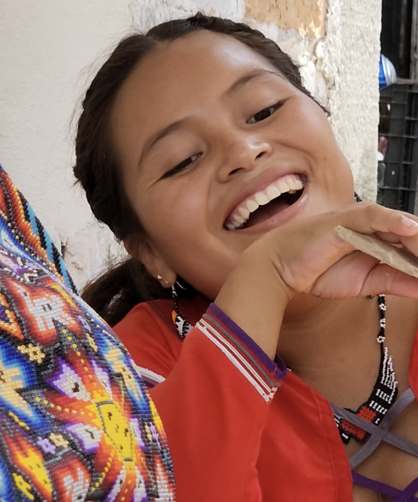 Primer plano de una niña huichol sonriente con vestimenta tradicional roja, luciendo aretes y collar de chaquira, con una colorida pieza de arte wixárika al fondo. Su expresión captura la alegría y el espíritu vivaz de la cultura huichol
