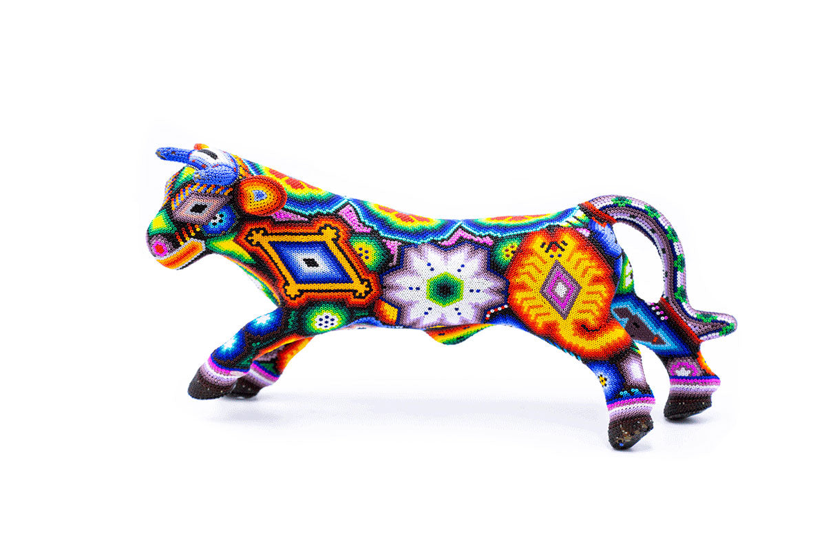 Escultura artesanal en forma de toro en plena carrera, meticulosamente decorada con un patrón de chaquiras vibrantes que forman intrincados diseños geométricos y florales en una variedad de colores brillantes como naranja, azul, verde y morado, sobre un fondo blanco puro