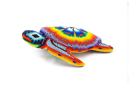 Escultura artesanal de una tortuga marina decorada con un mosaico de chaquira que representa patrones de arte Huichol en colores vibrantes como azul, rojo, amarillo y naranja. La pieza captura la forma natural de la tortuga con sus aletas extendidas, como si estuviera nadando